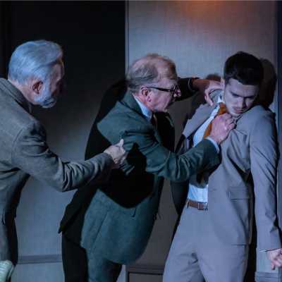 Three white men wearing suits. One is being pushed into the wall by another and the third is trying to hold one back. 