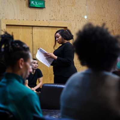 A person standing looking at a script is visible through blurry seated people 