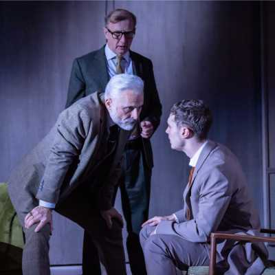 Three white men of average height wearing suits in a living room. A younger man with brown hair is sat in a chair; another man with silver hair and a beard leans over him intimidatingly with his hands on his knees, while the third man stands behind watching. 