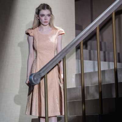 A petite, pale woman with chest-length blonde hair stands at the bottom of a staircase. She is looking at someone standing in the room at the bottom. She looks upset.