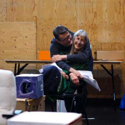 A smiling person sits on a chair in a rehearsal room with wooden walls. They are being hugged by a person stood behind them. 