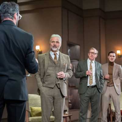 Four white men of average high in suits. One is facing away from the camera and the other three are looking at him. They are all holding glasses. 