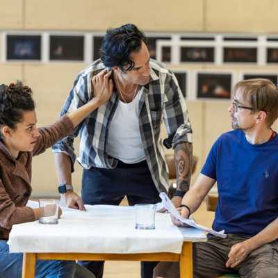 Ramin Karimloo, Anoushka Lucas and Howard Gossington sit around a table. Ramin is looking at Howard, unhappy about something he has said. Anoushka has her hand on Ramin, telling him to stop talking