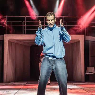 A young man in a pale blue tracksuit top stands centre-stage, gesturing with his hands forwards. Behind him, two people stand defiantly and watch. A tunnel is behind them, with a curved bridge over it, and there are red stage lights. 