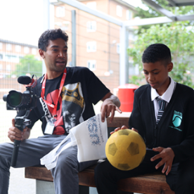 A person holding a camera on a gimble sits on a bench next to a student in a school uniform holding a football. 