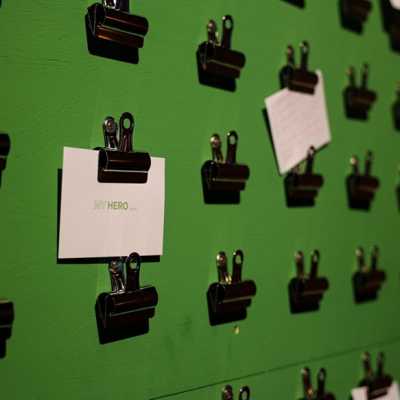 Rows of black bulldog clips stuck on a green wall. Multiple pieces of card are being held up by the clips out of focus. One is in focus that reads "My Hero..." 