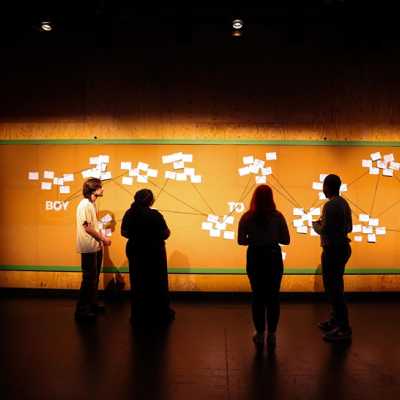 The backs of 4 people standing in front of a brown cork board with clusters of white cards with writing on them connected by twine. Words across the board left to right read "Boy to Man" 