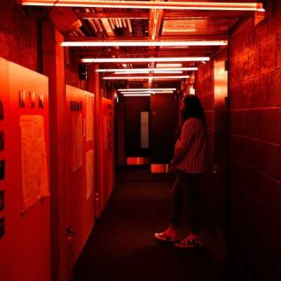 A corridor with orange lights along the ceiling. The left side of the corridor has orange walls with photos and A3 paper with writing stuck to them. A person in side profile is stood looking at the wall. 