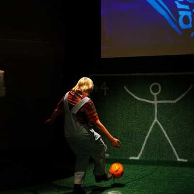 The back of a person kicking a football towards a grassy wall with a white stick figure drawn on it. 