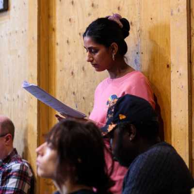 A person stands leaning against a wooden wall looking at a script. Blurred seated people are visible in the foreground. 