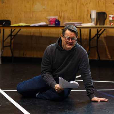 A person sits on the floor of a rehearsal room holding a script looking upset. 