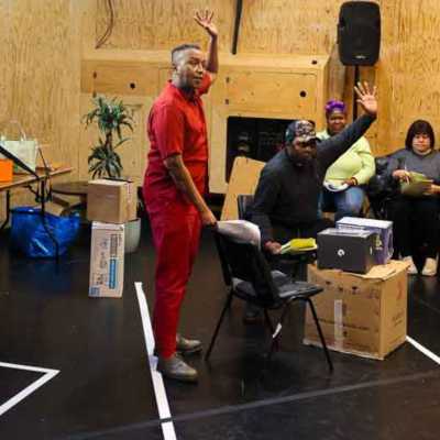 Two people , both with an arm raised, stand and sit in a rehearsal room. Two seated people are visible behind them.