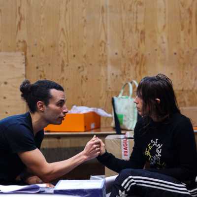 Two people sat facing each other in a rehearsal room with wooden walls. One person is leaned forward and gripping the other person's hand. 