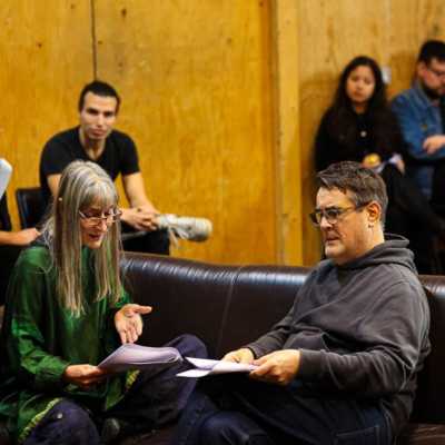 Two people sit of a sofa reading to each other from scripts. Blurred seated people are visible in the background. 