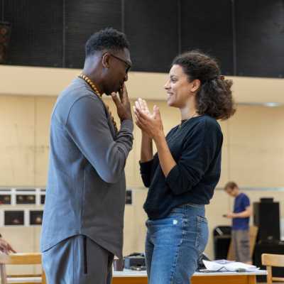 Director Kwame Kwei-Armah and lead actor Anoushka Lucas stand facing  eachother, in coversation. Other cast members can be seen in the background.