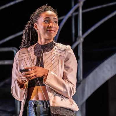 A woman, wearing a beige jacket, brown top, blue jeans and a sparkly handbag stands on a stage. She is holding a mobile phone in both hands and is looking to her left with a small smile on her face. Behind her is a metal bridge. 