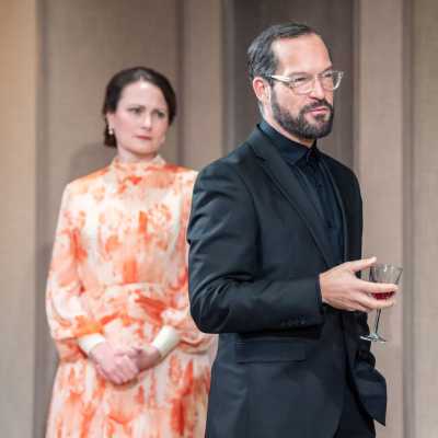 A white man of average height wearing a black suit and see through glasses. Behind him stands a tall woman with brown hair wearing a flowery orange and white dress. 