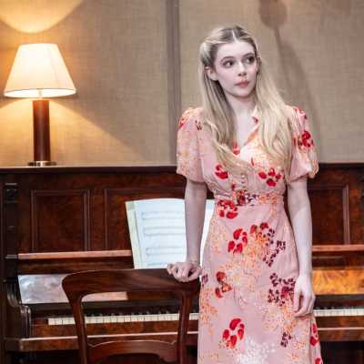 A petite, pale woman with chest-length blonde hair stands against a paino. She wears a pale pink dress with red, white and orange flowers on it. 