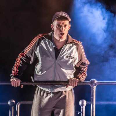 A young man wearing a grey Adidas tracksuit top, grey tracksuit bottoms and a Nike cap is leaning over a metal railing. His mouth is open, mid-speech, and he is wearing a serious expression.  