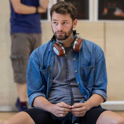 Solo shot of cast member Andrew Coshan. He is sitting down, looking at someone or something off camera. He wears wired headphones around his neck.