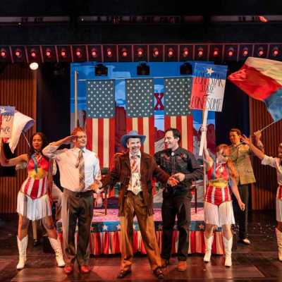 A group of people stand by a small float with USA flags on it. Some of them hold up signs. In the centre is a white man wearing a fringed jacket.