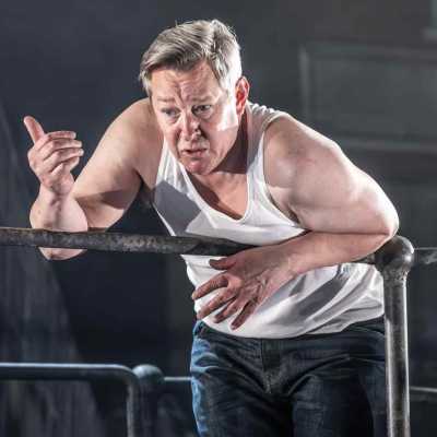 A man wearing a white vest and dark blue jeans is leaning over a metal railing. Both of his arms are resting over the rails, whilst his right hand is raised slightly. 