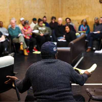 The back of a seated person with both arms out. One hand is holding a script. Behind them watching them are multiple blurred seated people. 