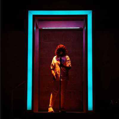 The silhouette of a woman holding a microphone stands in the middle of a pink and blue illuminated doorway.