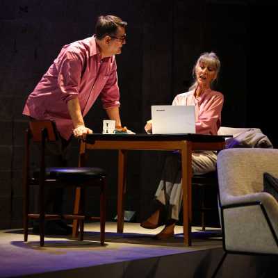 A white woman in a pink shirt, sits at a coffee table looking at her laptop, looming over her is a white man also in a pink shirt, wearing glasses who looks directly at her.