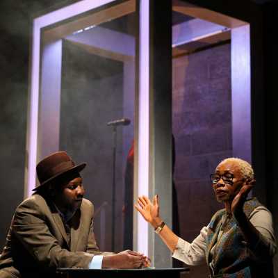 A black man in a suit and fedora sits at a table in conversation with a black woman with short blonde hair, her hands are held up in a defeated way, behind them is a futuristic looking frame with a person centred inside it.