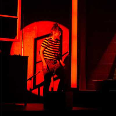  A woman stands slightly bent over, the stage is illuminated by red light and her top half is spotlighted by a sinister red circle, she is wailing. 