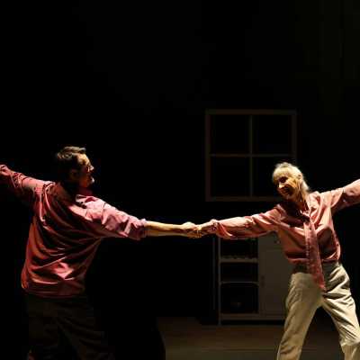 A man and woman both wearing pink shirts, gleefully look at each other, they are dancing, each clasping hands with one arm and extending the other arm outwards. 