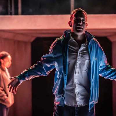 A man, wearing a blue tracksuit top, white shirt and blue jeans stands on a stage. His mouth is slightly open, face slightly hidden behind shadows from the stage lights, and his arms are out to the sides. Behind him on the left, out of focus, is a woman wearing a hoodie and jeans.  