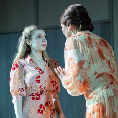 Two women wearing dresses with orange and red tones are having a conversation. One with brown hair, stands facing away from the camera and is saying something. The other has chest-length blonde hair. 