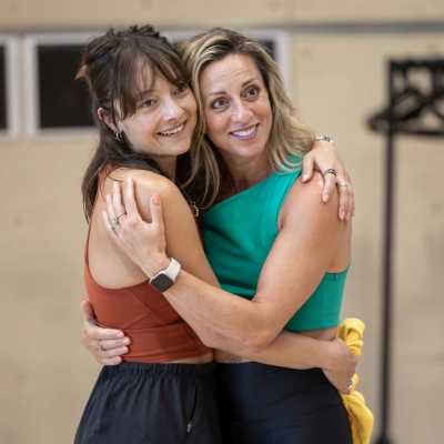 Company member Jasmin Colangelo and Choreographer Lizzi Gee are hugging eachother and smiling. Jasmin is wearing a brown top and black tracksuit bottoms. Lizzi is wearing a green top and balck leggings.