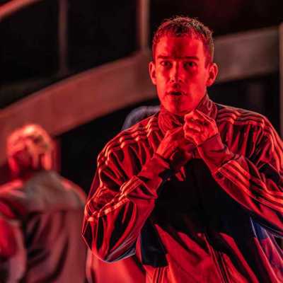 A half-body shot of a man wearing an Adidas tracksuit top. He is staring intensely, with his mouth slightly open. His hands are on the zip of his top, as if doing it up. The image has a red hue from the stage lighting.