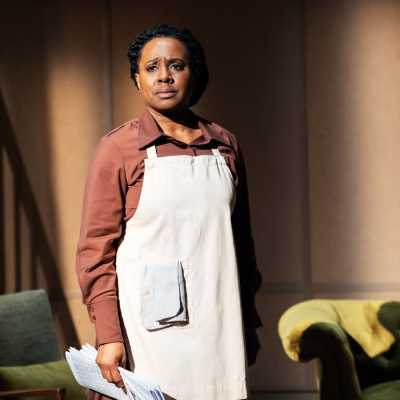 A Black woman with dreadlocks that are tied back, faces the camera. She wears a brown dress with a white apron over the top of it and carries papers in one hand. 