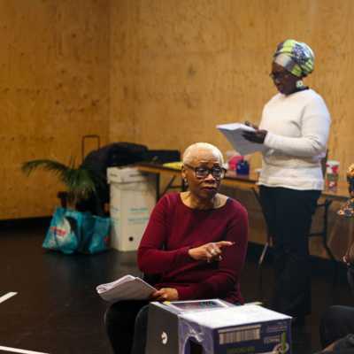 Two people holding scripts acting in a rehearsal room. Two are seated at a makeshift table speaking to each other; the third stands behind them.