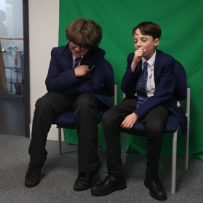 Two students sit in chairs in front of a green screen performing. 
