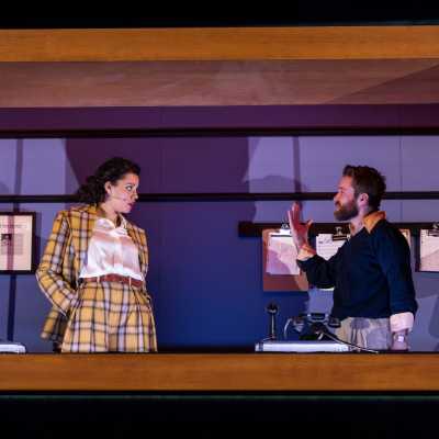 A small office room bordered with wood. Inside is a light-skinned mixed race woman wearing yellow, and a white man with light brown hair and a beard.