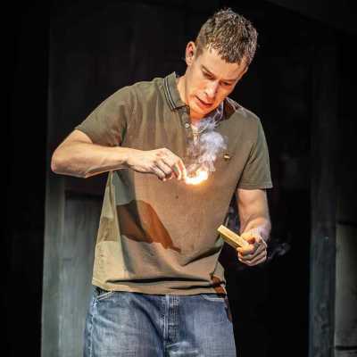 A man in a green polo shirt and blue jeans stands behind a metal bin. He is holding a matchbox in his left hand and is staring intensely at the lit match in his right hand. Smoke from the match is going upwards towards his face.  