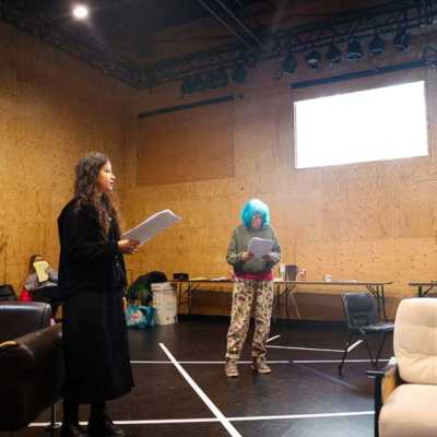 Two people stand holding scripts and speaking to each other in a rehearsal room with a sofa, armchair and wooden walls. 