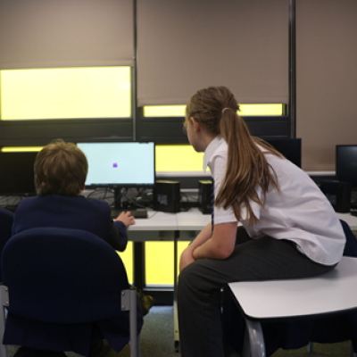 One student sits on a chair working at a computer while another student sits on the desk next to them looking at the screen. 