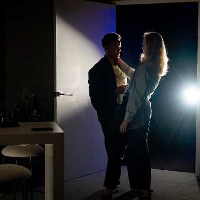 Two people stand in the entrance to a modern apartment lit from behind: one brown-skinned, non-binary woman with short Afro hair, and one a white Welsh woman with blonde shoulder-length hair. 