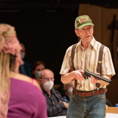 Greg Hicks, as Andrew Carnes pointing gun, in focus towards another character on stage with Marisha Wallace as Ado Annie stood diagonal from him, with her back to the camera