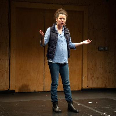 A woman stands in denim with her hands wide, speaking to the audience.