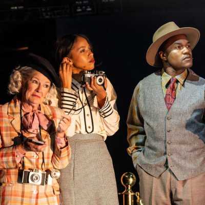 Three people stand, holding cameras and mics: a woman in her 60s, who is quite short, a tall mixed-race woman and a small Afro-Caribbean man.