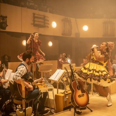 Marisha Wallace as Ado Annie sings by the band, opposite Liza Sadovy as Aunt Eller who is also singing, they are surrounded by a hazy, golden light