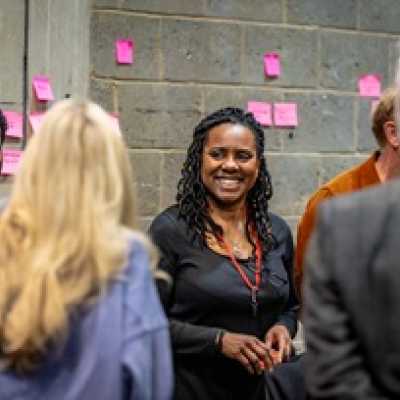 A group of people stand in a circle. A Black woman with shoulder length dreadlocks is facing the camera, smiling. 