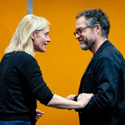 A blonde woman and a white man with brown hair are talking. Behind them is a bright orange background. She is touching his arm and they are smiling as if sharing a joke. 